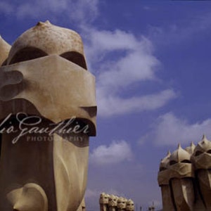May include: A close-up view of several unique, sculpted, beige-colored chimney tops against a blue sky with white clouds. The chimney tops are shaped like human figures with rounded heads and geometric features.