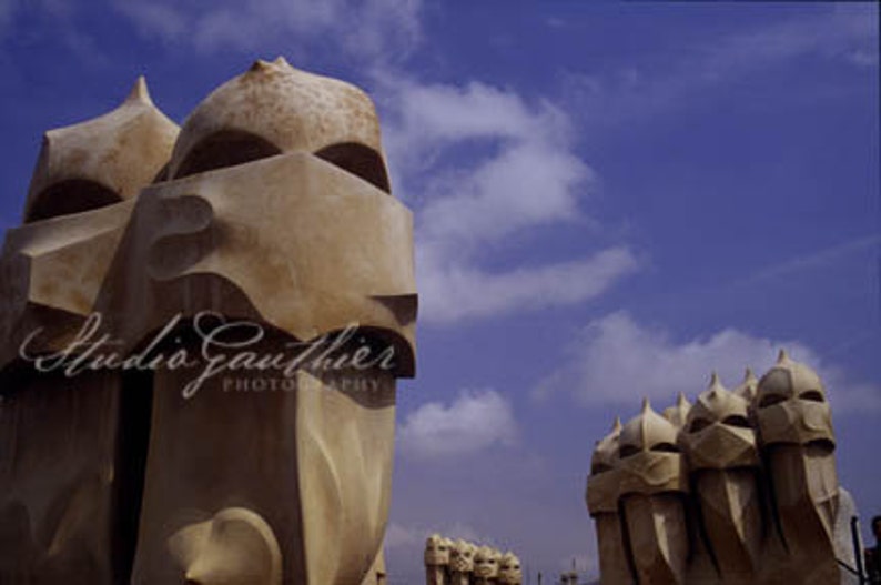May include: A close-up view of several unique, sculpted, beige-colored chimney tops against a blue sky with white clouds. The chimney tops are shaped like human figures with rounded heads and geometric features.