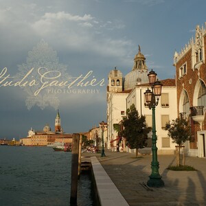 May include: A view of a canal in Venice, Italy, with a large building on the right side of the image. The building has a red brick facade and a series of arched windows. There is a street lamp in the foreground, and a body of water in the background.