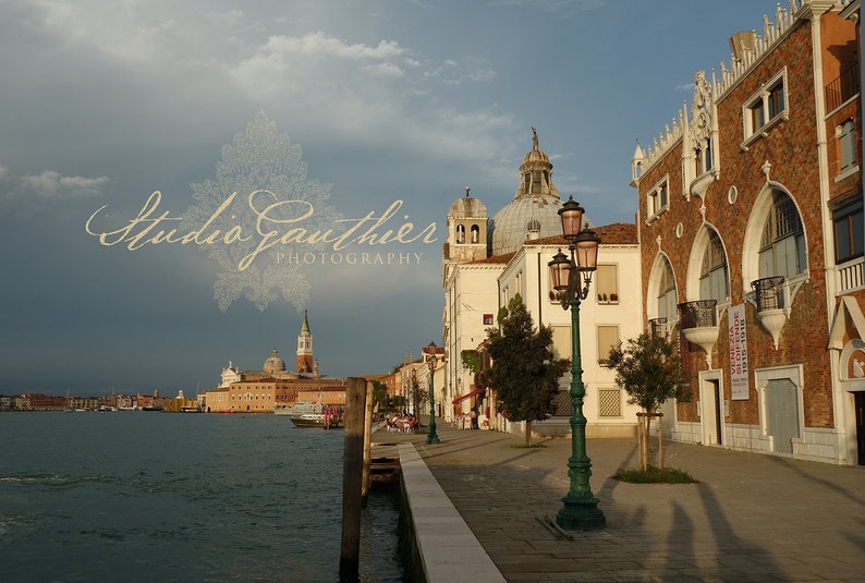 May include: A view of a canal in Venice, Italy, with a large building on the right side of the image. The building has a red brick facade and a series of arched windows. There is a street lamp in the foreground, and a body of water in the background.