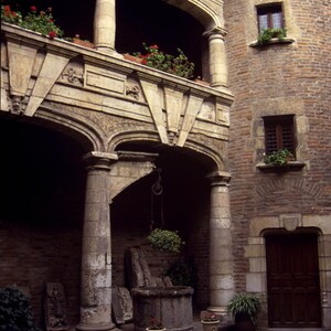 May include: A stone courtyard with a well in the center, surrounded by arches and columns. The courtyard is part of an old building with brick walls and windows.