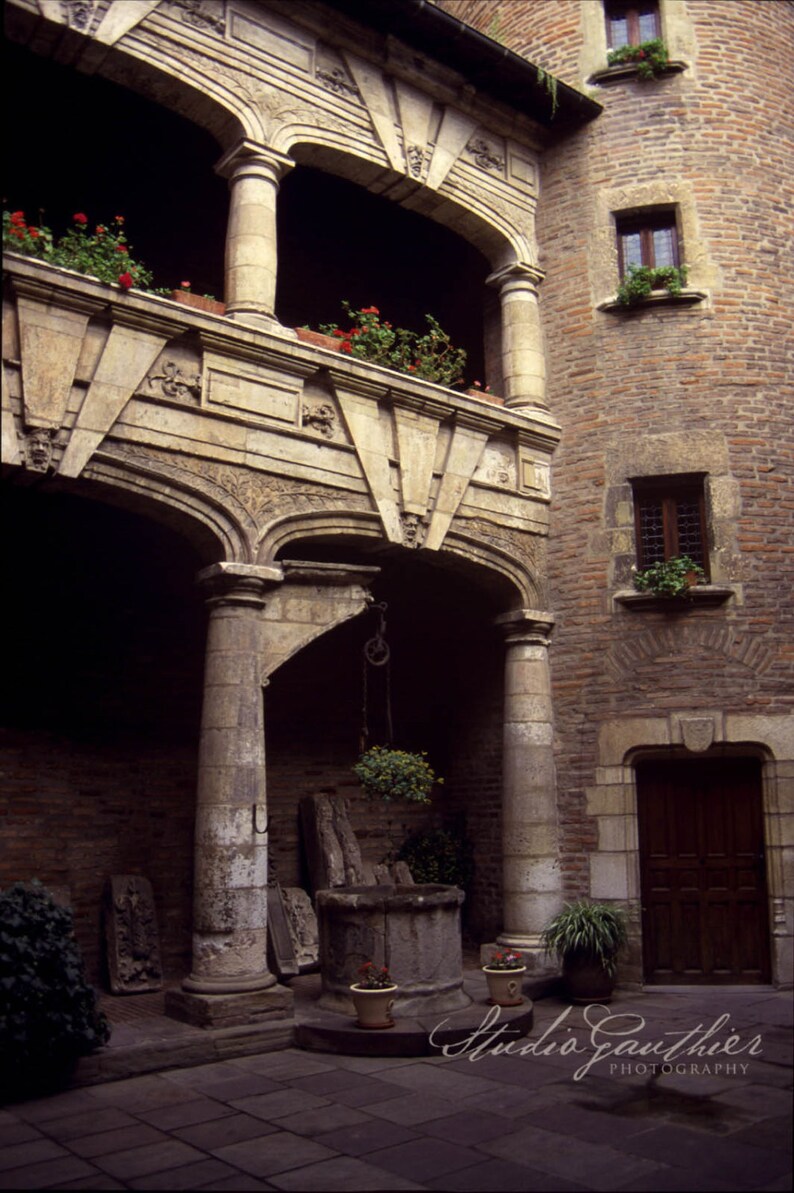 May include: A stone courtyard with a well in the center, surrounded by arches and columns. The courtyard is part of an old building with brick walls and windows.