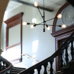 May include: A black metal chandelier with multiple light bulbs hangs from the ceiling of a room with a wooden staircase. The staircase has a black railing and a white wall behind it.