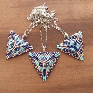 Puede incluir: Tres colgantes triangulares con cuentas y cadenas de plata. Los colgantes presentan un patrón geométrico en tonos azul marino, azul claro y beige. Los collares se exhiben sobre una superficie de madera marrón.