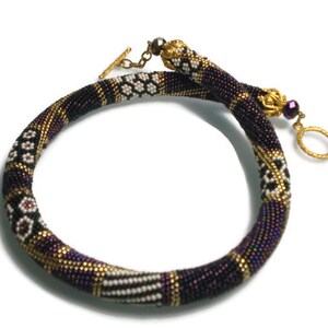 May include: A beaded necklace in shades of purple, black, and gold. The necklace features intricate patterns and a gold-tone clasp. The design includes white and black geometric shapes. The necklace is approximately 16 inches in length.