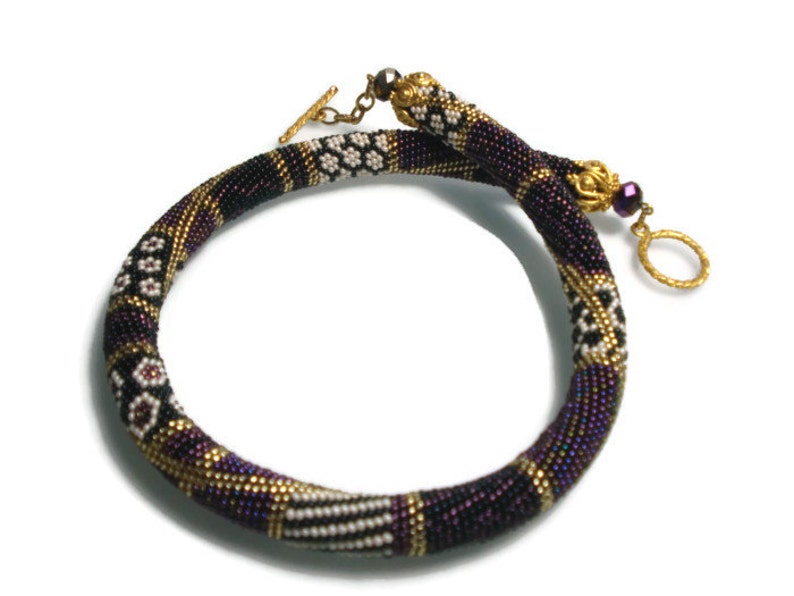 May include: A beaded necklace in shades of purple, black, and gold. The necklace features intricate patterns and a gold-tone clasp. The design includes white and black geometric shapes. The necklace is approximately 16 inches in length.