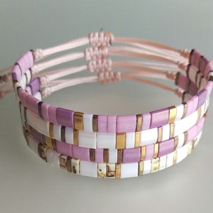 Könnte beinhalten: Ein mehrlagiges Armband mit rosa, weißen und goldenen Perlen. Das Armband ist mit einer geknoteten Schnur gefertigt und hat einen Quasten.