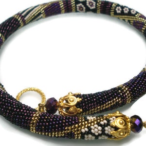 May include: A beaded necklace in shades of purple, black, gold, and white. The necklace features a tubular design with intricate beadwork and gold-colored metal accents. The clasp is a gold-colored toggle. The necklace is approximately 16 inches long.