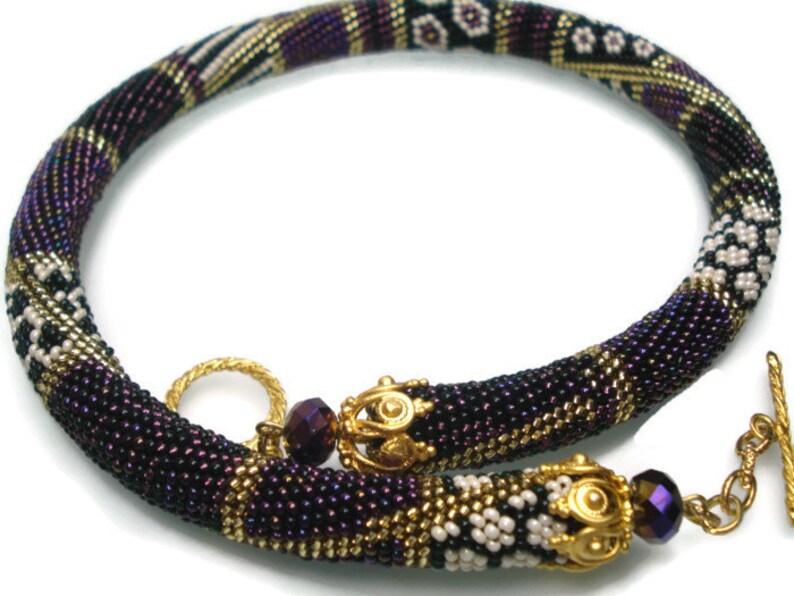 May include: A beaded necklace in shades of purple, black, gold, and white. The necklace features a tubular design with intricate beadwork and gold-colored metal accents. The clasp is a gold-colored toggle. The necklace is approximately 16 inches long.