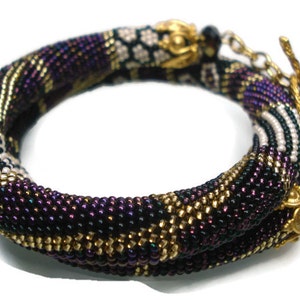 May include: A beaded bracelet in shades of purple, black, gold, and white. The bracelet is made of small beads woven together in a spiral pattern. The clasp is gold-colored, and the bracelet is designed to wrap around the wrist.