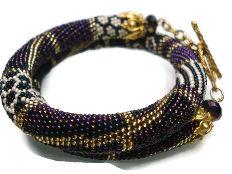 May include: A beaded bracelet in shades of purple, black, gold, and white. The bracelet is made of small beads woven together in a spiral pattern. The clasp is gold-colored, and the bracelet is designed to wrap around the wrist.