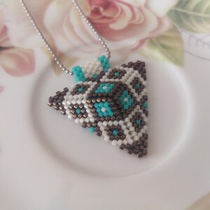 Op de afbeelding: Een ketting met een driehoekige hanger met een geometrisch patroon van witte, bruine en turquoise kralen.
