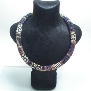 May include: A beaded necklace displayed on a black bust. The necklace features a cylindrical design with a mix of colors, including black, purple, gold, and white. The beads are arranged in a repeating pattern, creating a textured appearance. The necklace is a statement piece.