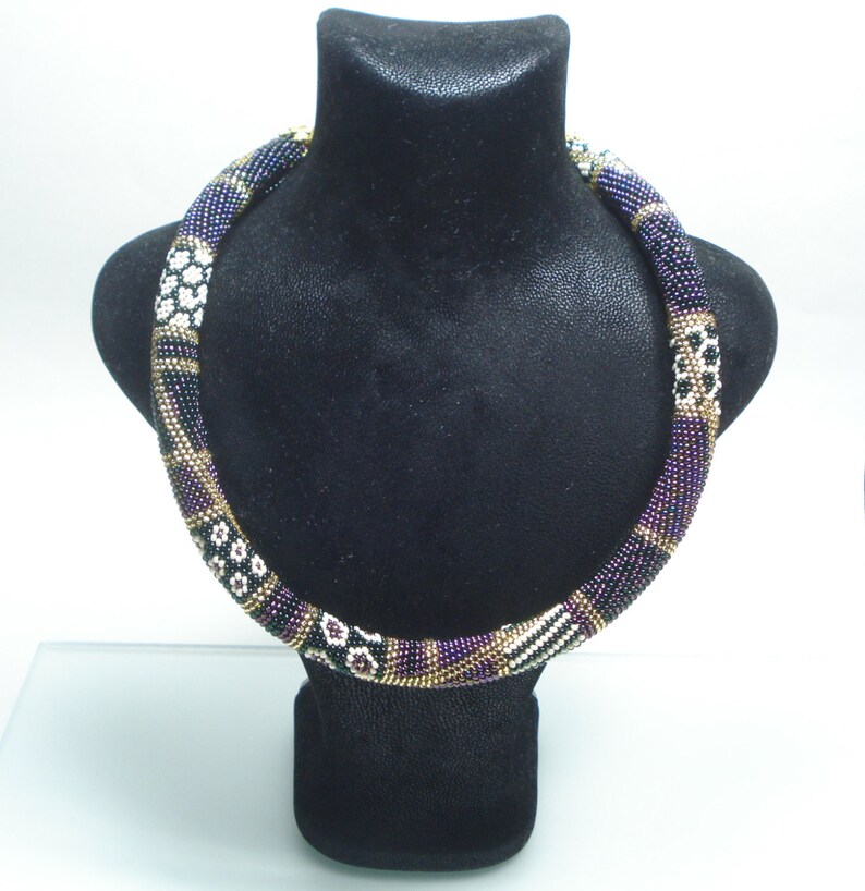May include: A beaded necklace displayed on a black bust. The necklace features a cylindrical design with a mix of colors, including black, purple, gold, and white. The beads are arranged in a repeating pattern, creating a textured appearance. The necklace is a statement piece.