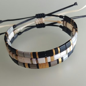 Op de afbeelding: Twee gelaagde armbanden met zwarte, witte, gouden en grijze vierkante kralen. De armbanden zijn gebonden met zwarte en witte koorden.