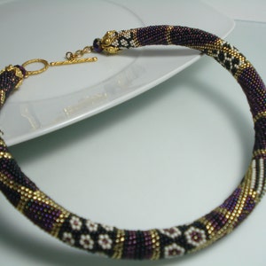 May include: A beaded necklace with a gold clasp. The necklace features a pattern of black, purple, white, and gold beads. The necklace is circular and appears to be handmade.