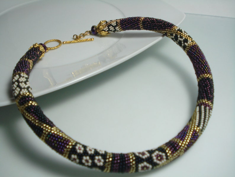 May include: A beaded necklace with a gold clasp. The necklace features a pattern of black, purple, white, and gold beads. The necklace is circular and appears to be handmade.