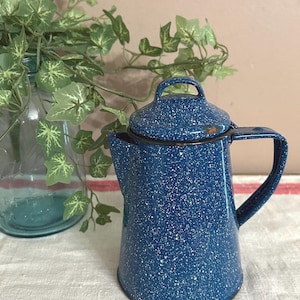 May include: A blue enamelware coffee pot with a lid and handle. The pot has a speckled white pattern and a black rim. A glass jar with greenery is in the background.