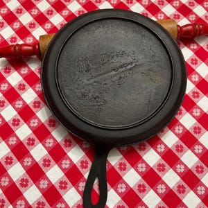 May include: A black cast iron griddle with a long handle and a wooden rolling pin with red handles. The griddle is round and has a slightly textured surface. The rolling pin is resting on top of the griddle.