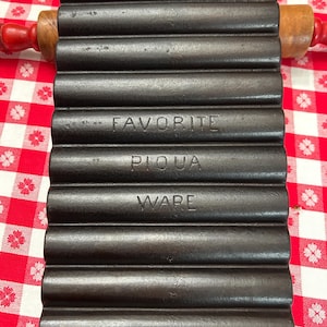 May include: A cast iron cornbread mold with the words "FAVORITE PIQUA WARE" embossed on the front. The mold has a series of parallel, rounded troughs. A wooden rolling pin with red handles rests on the mold.