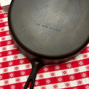 May include: A round black cast iron skillet with a handle featuring a hole at the end. The underside of the skillet is embossed with the text 14 INCH SKILLET and a small letter A. The surface of the metal is dark, textured, and matte.