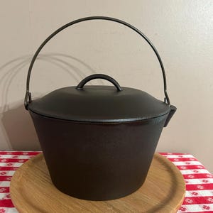 May include: A dark brown cast iron pot with a matching lid and a curved metal bail handle. The pot features a tapered, cylindrical body with a smooth finish and a small pouring spout on the side, designed for cooking and food preparation.