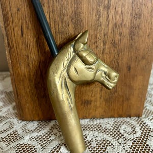 May include: A brass horse head handle attached to a thin black rod. The horse head features detailed molded facial features, including ears, eyes, and a mane, with a polished metallic finish. The rod extends upward from the top of the horse head.