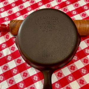 May include: A dark brown cast iron skillet with a round base and a handle. The underside features embossed text reading WAGNER WARE SIDNEY -O- and the number 1053N. The skillet is positioned over a wooden rolling pin with red handles.