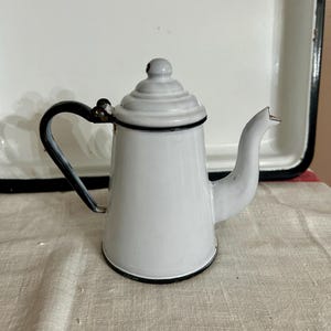 May include: A small, white enamelware teapot with a black handle and trim. The lid is also white and has a small knob on top. The teapot is sitting on a beige surface, with a white tray in the background.