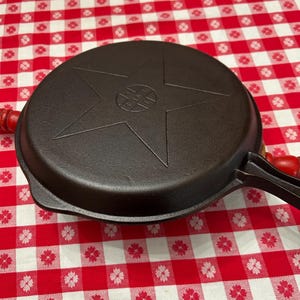 May include: A black cast iron skillet with a star design on the lid. The skillet has a handle with a red wooden grip. The skillet is sitting on a red and white checkered tablecloth.
