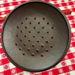 May include: A dark brown, cast iron lid with a series of evenly spaced, rounded bumps across its surface. The lid has a circular shape with a slightly raised edge. The number "8" is visible on the lid. The lid is on a red and white checkered surface.