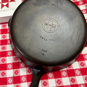 May include: A round black cast iron skillet with a handle. The underside features the embossed text NO. 8, a circular logo with the word GRISWOLD inside, the text ERIE PA., and the numbers 704 followed by the letter R below the logo.