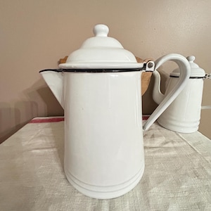 May include: White enamel coffee pot with a black rim and handle. The pot has a lid with a small knob and a spout for pouring. A smaller white pot is visible in the background.