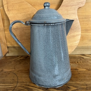 May include: A vintage gray enamelware coffee pot with a lid and handle. The pot has a speckled pattern and a spout for pouring. It is sitting on a wooden surface with a wooden chicken-shaped cutting board in the background.