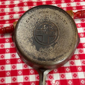May include: A well-worn cast iron skillet with the text "CAST IRON SKILLET" and "GRISWOLD ERIE PA U.S.A." The skillet is dark gray with a handle and is resting on a red and white checkered surface.