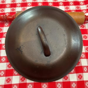 May include: A dark brown cast iron skillet lid with a wooden handle. The lid is round and has a slightly domed shape. The handle is a dark brown color. The lid is sitting on a red and white checkered tablecloth.