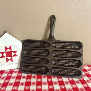 May include: A dark brown cast iron corn stick pan featuring eight individual corn-shaped molds arranged in two columns of four. The pan has a single handle at the top center. Next to it is a white house-shaped sign with a red star pattern.