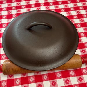 May include: A dark brown cast iron pot lid with a curved handle. A wooden rolling pin with red handles rests beneath the lid. The items are on a red and white checkered tablecloth.