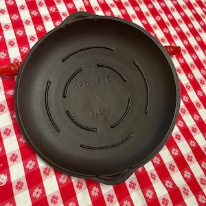 May include: A dark brown cast iron skillet with the text "IRON AGE No 8". The skillet has a circular design with curved lines and red handles. It sits on a red and white checkered tablecloth.