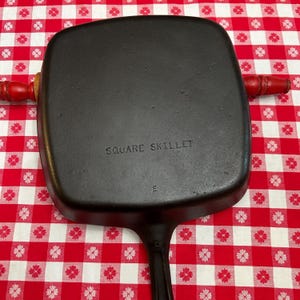 May include: A black cast iron square skillet with a handle, featuring the embossed text SQUARE SKILLET and the letter E on the underside. The skillet is positioned over a red and white checkered rolling pin with red wooden handles.