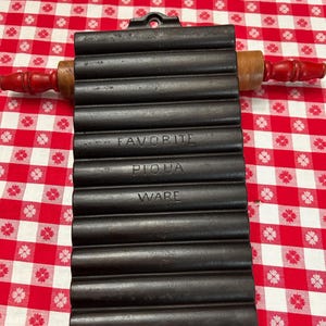 May include: A black cast iron bread mold with the words "FAVORITE PIQUA WARE" embossed on the front. A wooden rolling pin with red handles rests on top of the mold. The mold is on a red and white checkered tablecloth.