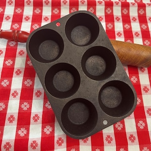 May include: A dark gray cast iron muffin pan with six circular indentations arranged in two rows of three. The pan features a small circular hole at the top and bottom center for hanging. The surface of the metal has a textured, matte finish.