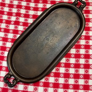 May include: An oval-shaped dark brown metal tray with a smooth, flat center and a raised rim. The tray features two decorative, ornate handles on opposite ends, each with a cutout design. The surface shows a weathered, aged patina throughout.