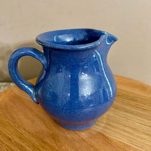 May include: A small, rounded ceramic pitcher with a vibrant, deep blue glaze and a glossy finish. The pitcher features a curved handle on the left side, a wide opening at the top, and a small, integrated pouring spout on the right side.