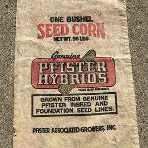 May include: A beige fabric sack featuring black and red text that reads ONE BUSHEL SEED CORN NET WT. 56 LBS. GENUINE PFISTER HYBRIDS, and GROWN FROM GENUINE PFISTER INBRED AND FOUNDATION SEED LINES. PFISTER ASSOCIATED GROWERS, INC. includes a corn cob graphic.