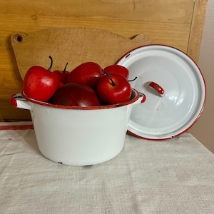 May include: A white enamel pot with red trim and handles, filled with red apples. A matching lid with a red handle sits nearby. The pot is on a white tablecloth, with a wooden cutting board in the background.