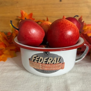May include: A white enamel bowl with a red rim and a handle, featuring a vintage Federal Vogue Enameled Ware logo. The bowl is filled with several bright red artificial apples, and the logo includes the text Federal Vogue Enameled Ware.