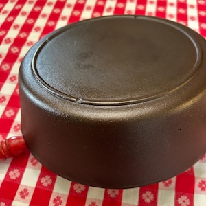May include: A dark brown cast iron skillet with a rounded base and a circular cooking surface. The skillet has a raised rim and a smooth, textured finish. A small, red wooden handle is visible on the side. The skillet is sitting on a red and white checkered tablecloth.