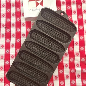 May include: A dark brown cast iron corn stick pan featuring seven individual molds shaped like ears of corn. The rectangular pan has a handle at each end and is positioned next to a white wooden block with a red star pattern and the text A String.