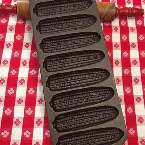May include: A dark brown cast iron corn stick pan featuring eight individual molds shaped like ears of corn, arranged in two columns. The pan has a rounded rectangular shape with a small hole at the top and bottom for hanging storage.